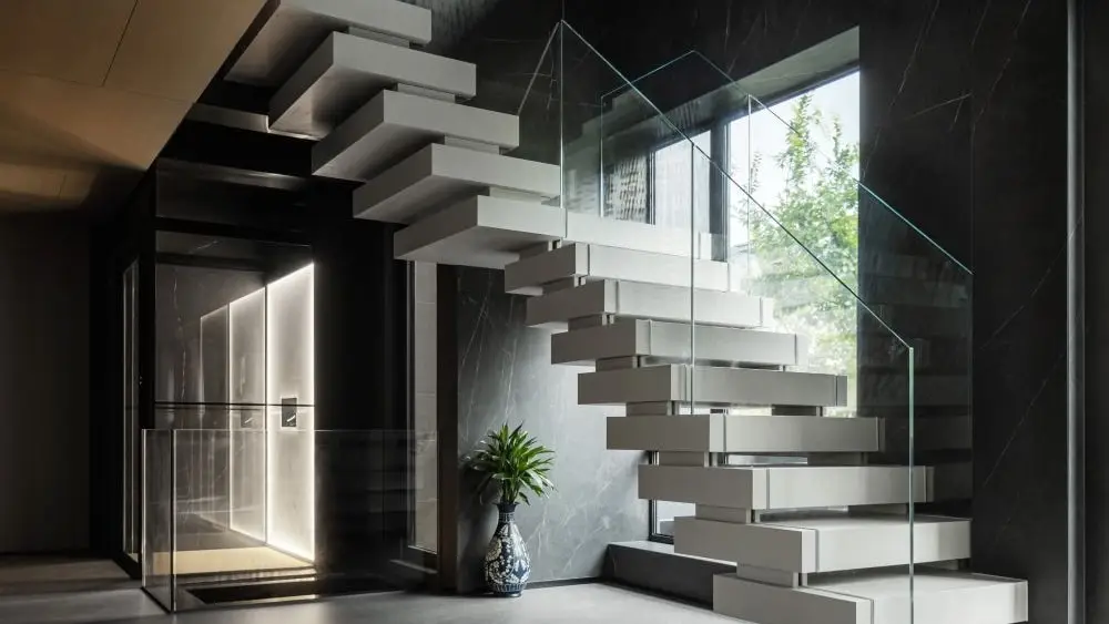 A glass-fronted platform lift installed beside a cantilevered staircase in a dark marble hallway.